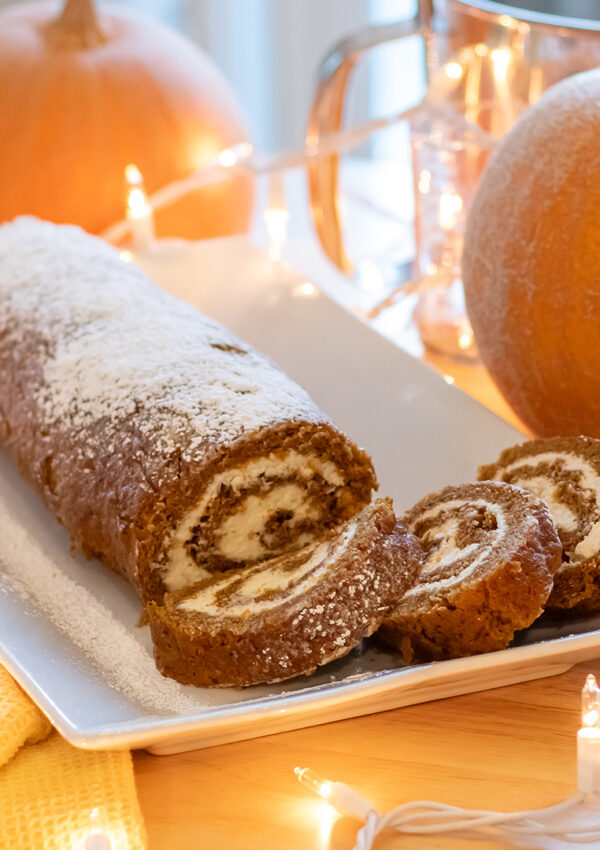 A pumpkin swiss roll, a slice cut to show the creamy swrirl of filling on the inside, searved on a dish. The roll is decorated by pumpkins and white decorative Christmas lights.