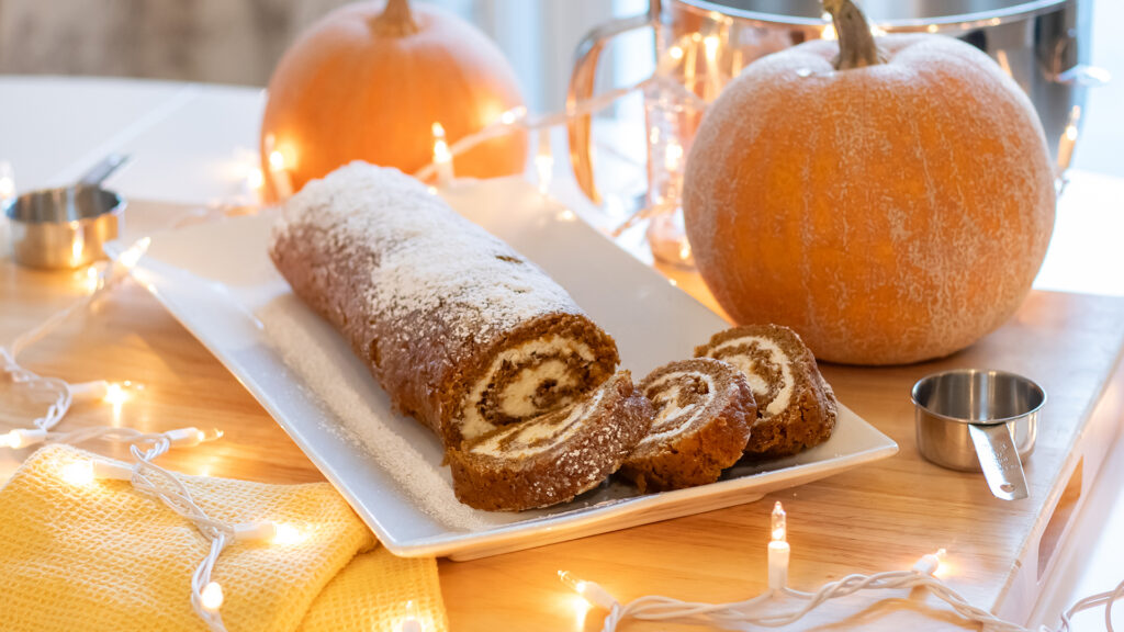 A photo of a pumpkin swiss roll, a slice cut to show the creamy swrirl of filling on the inside, searved on a dish. The roll is decorated by pumpkins and white decorative Christmas lights.