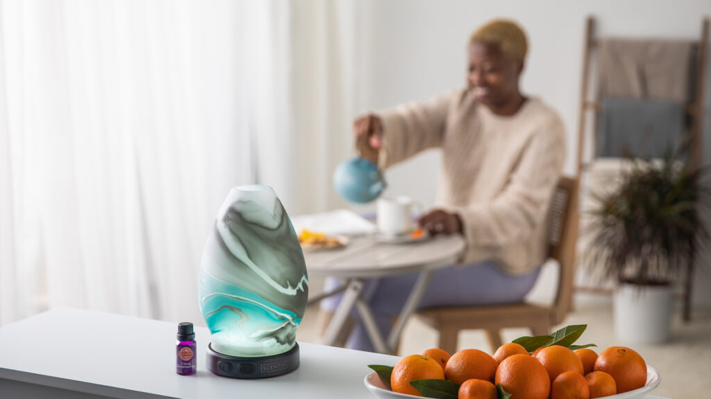 An image of a Scentsy Diffuser won a kitchen countertop next to a bowl of oranges. A woman pours a mug of tea in the background.