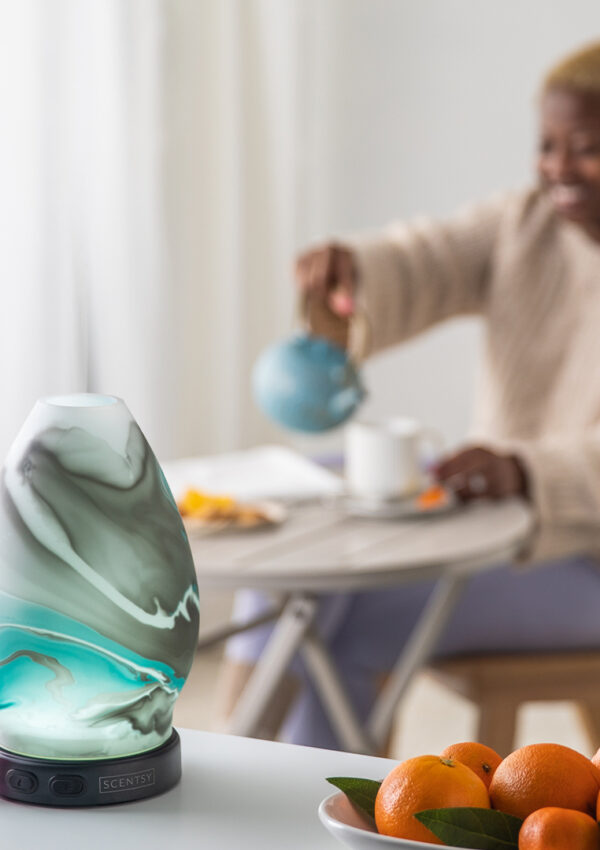 An image of a Scentsy Diffuser won a kitchen countertop next to a bowl of oranges. A woman pours a mug of tea in the background.