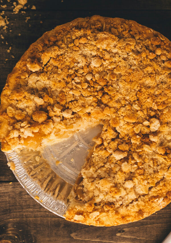 A golden-crusted apple pie with a slice missing sits on a table beside cut apples, butter, and a knife.