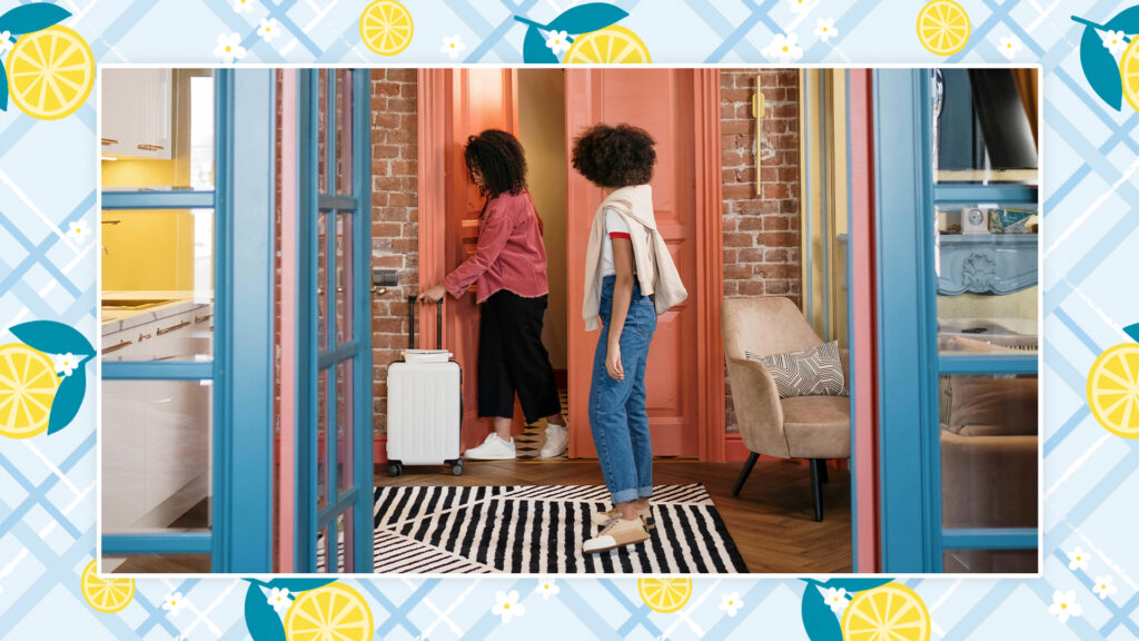 Two women with travel luggage stand in the entranceway of a vacation home.