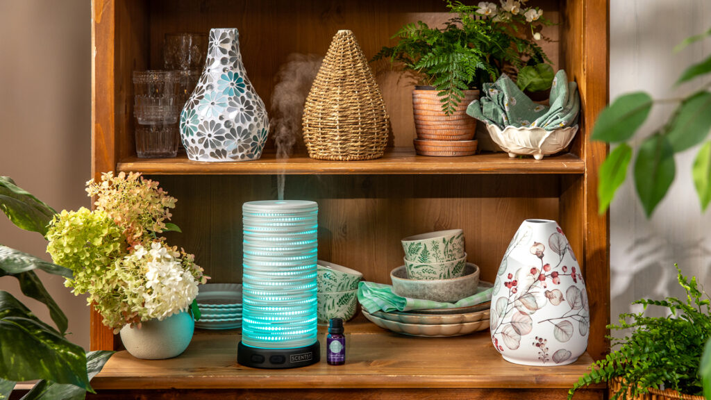 A wooden shelf with various Scentsy diffuser shades with floral and wicker patterns, along side dishes, flowers, and house plants. 