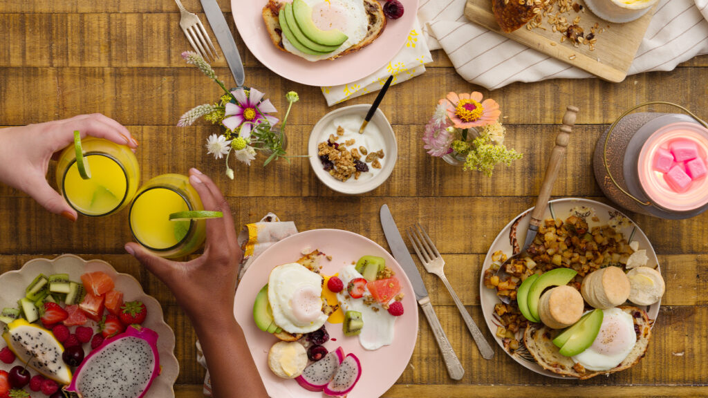 A summary breakfast of fruits, eggs, avocado, and fruit juice is served on pink plates with Scentsy fragrance flowers and and a warmer as decor among the dishes and food.