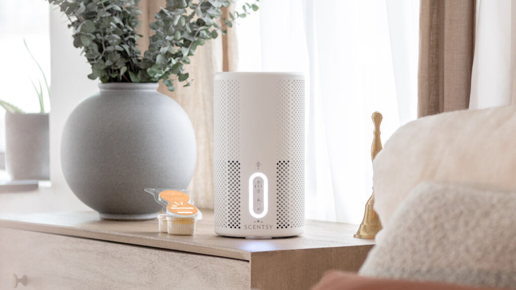 A white Scentsy Air Purifier sits on a beige bedside table beside a house plant and a Scentsy Pods Twin Pack.