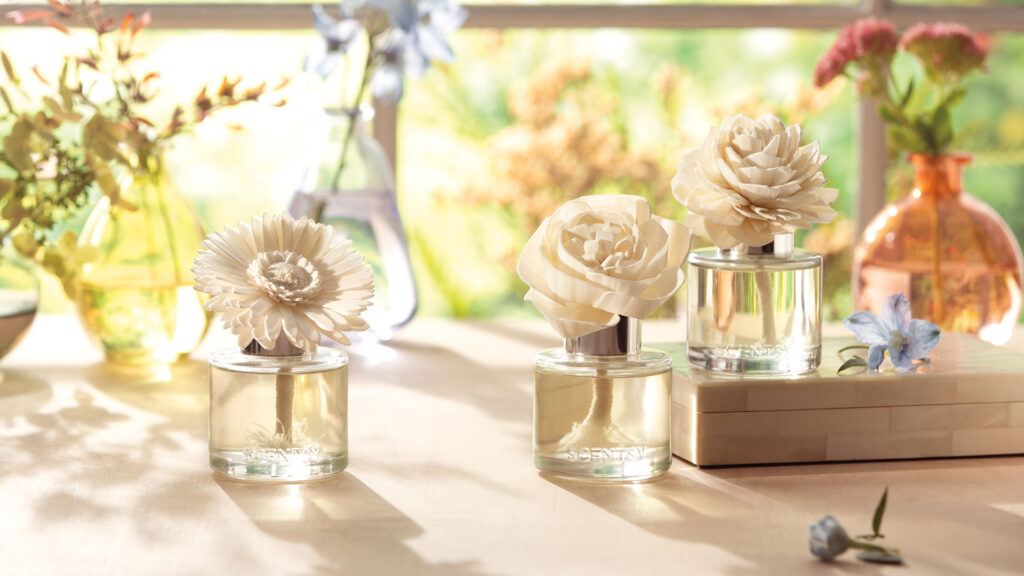Three Scentsy Fragrance flowers sit in a windowsill, with the sun shining brightly on the garden in the background.