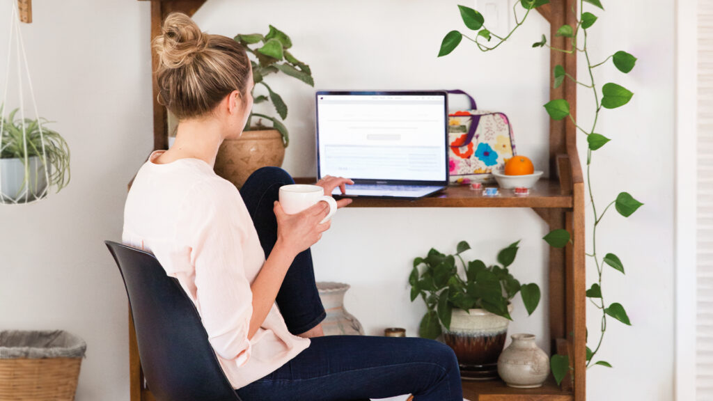 Woman in a white shirt sits sipping coffee in front of a laptop. She studies on the computer at a wooden desk and shelf covered with house plant decorations.