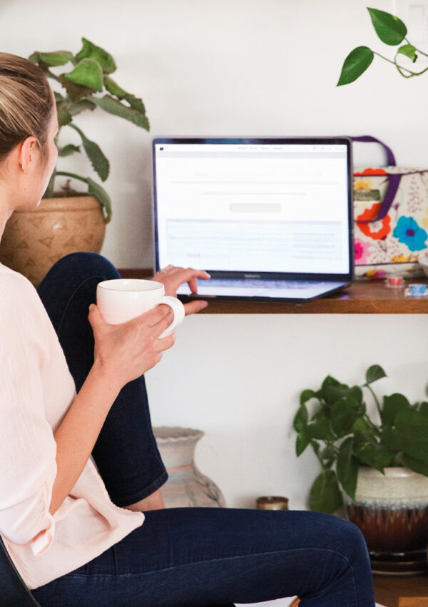 Woman in a white shirt sits sipping coffee in front of a laptop. She studies on the computer at a wooden desk and shelf covered with house plant decorations.