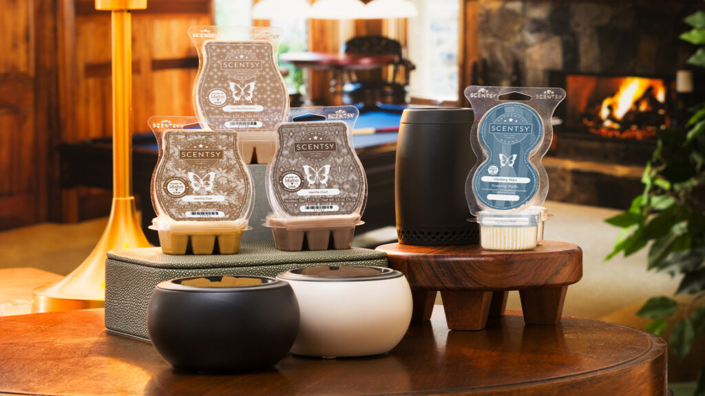 In a warmly lit room with a fireplace in the background, three Scentsy Wax Bars from the Father's Day Collection are displayed beside black and white Scentsy Mod Warmers. A Scentsy Go sits on a small wooden pedistal beside a plastic pack of Scensty Pods.