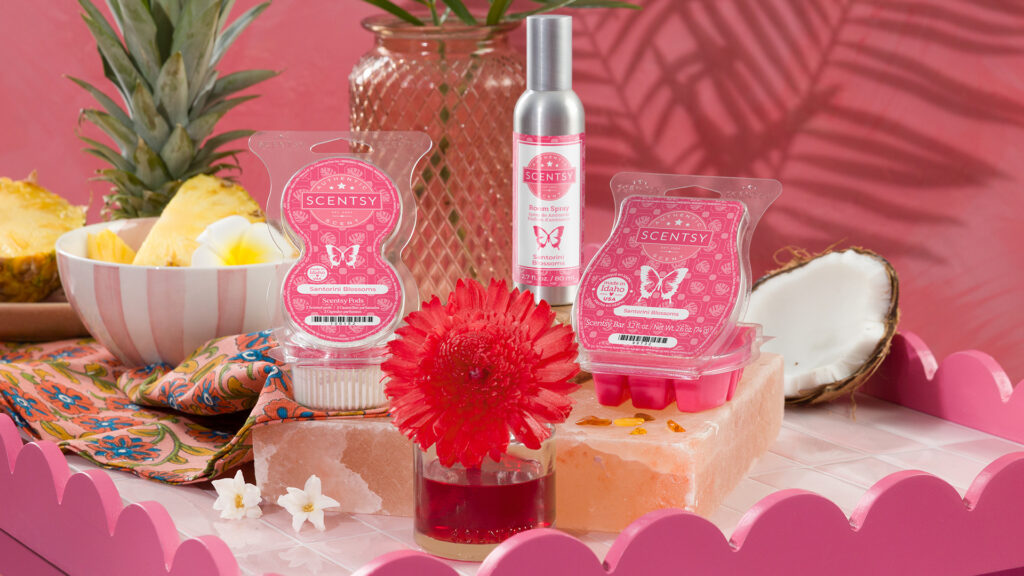 A Scentsy Fragrance Flower, Wax Bar, Room Spray, and Scentsy Pod Twin Pack, all in Santorini Blossoms fragrance, are staged on a pink-themed set with soaps, pink bowls, blossoms, and pineapples.