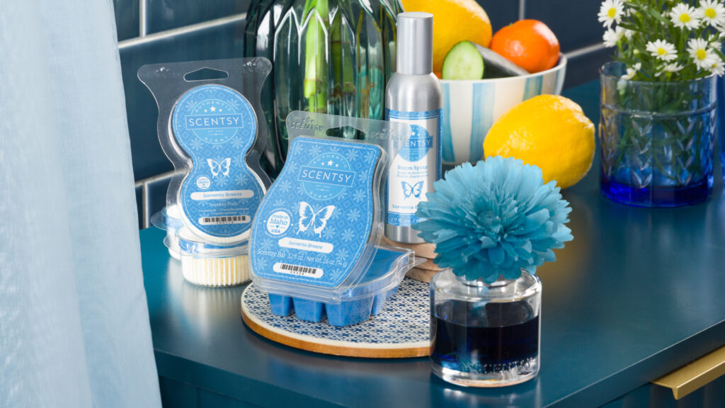 A Scentsy Fragrance Flower, Wax Bar, Room Spray, and Scentsy Pod Twin Pack, all in Sorrento Breeze fragrance and with blue lables. They sit on a blue countertop with blue tile and a blue bowl containing assorted fruits.
