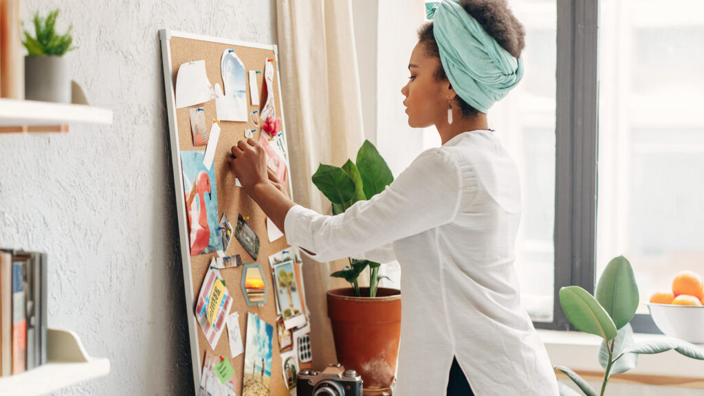 A black woman in a white shirt and blue hair wrap pins a photo onto a collage memory board.