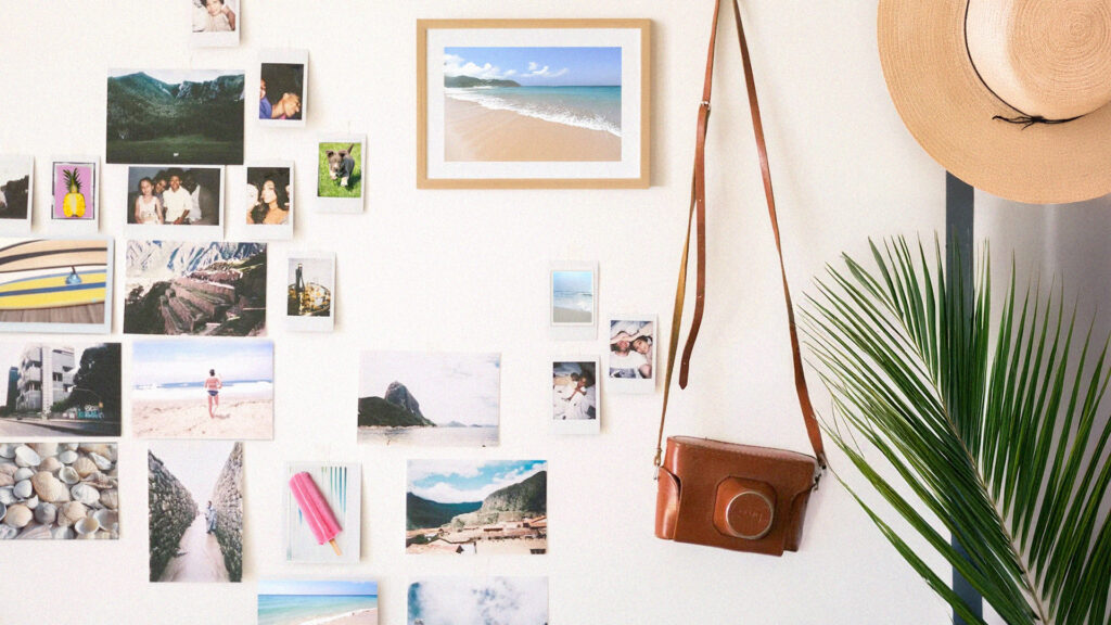 Dozens of photos depicting candid summer memories and beach landscapes cover a beige wall. From a hook hangs a camera in a leather case hangs, with a straw hat and palm frond beside it.