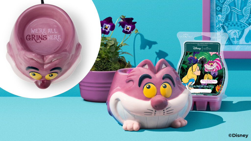 The Disney Cheshire Cat: Warm Fuzzies - Scentsy Warmer with the Disney We're All Mad Here Scentsy Wax bar beside it, both sitting on a blue table with a blue backgroud and purple blossomed pansies in a purple pot beside them.