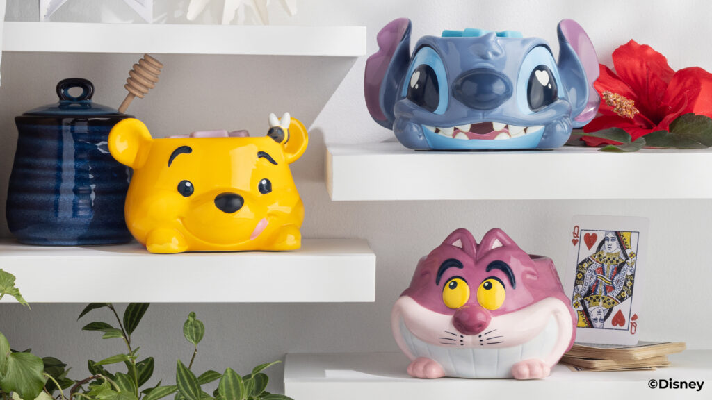 The three Disney Warm Fuzzy Warmers are displayed on tiered white shelves. The Stitch Warmer is posed on the top shelf beside a red hibiscus blossom. The Winnie the Pooh warmer is posed in the middle beside a honey pot. The Cheshire Cat Warmer is posed on the bottom shelf beside a queen of hearts playing card.