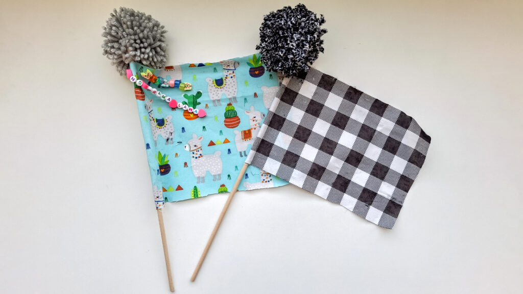 Two home mad po-topped flags lay side by side on a white table. One flag is light blue with llamas and cactus decorations on it. The other is black, white, and grey checked. Both flags are topped with grey and black pom poms.