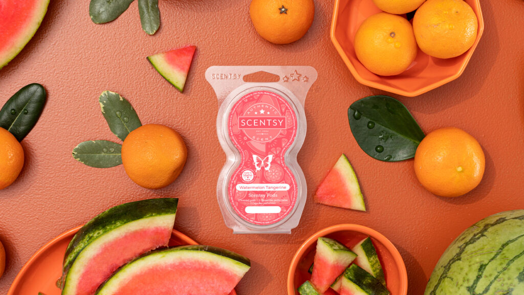 A single Scentsy Bod in Watermelon Tangerine fragrance sits on a tangerine colored background surrounded by dark green leaves, sliced watermelon wedges, and orange tangerines.