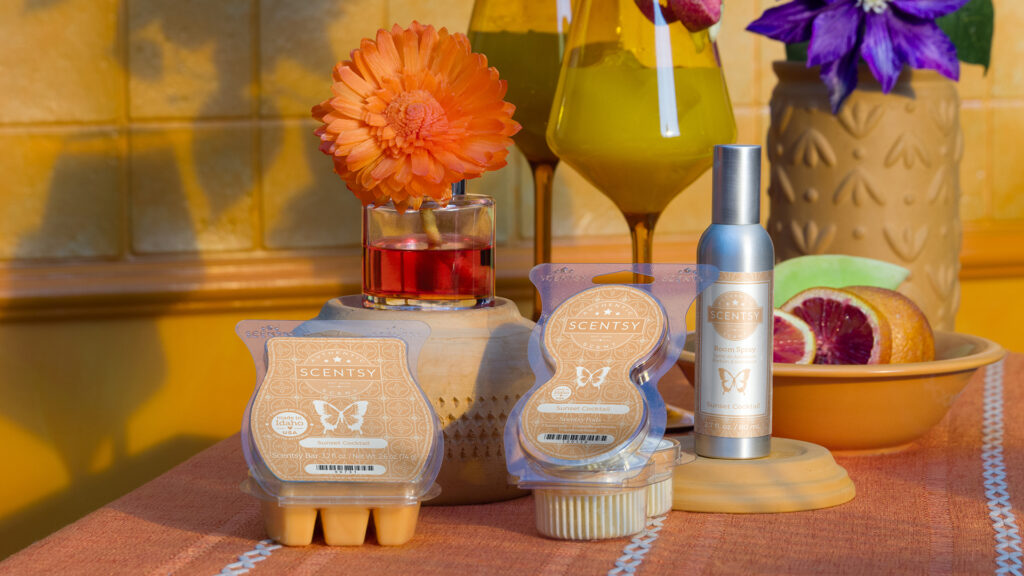 Scentsy Sunset Cocktail Wax Bar, Fragrance Flower, Room Spray, and Pods decorate a tabletop with glasses, vases, and bowls, all in a warm sand and brick color scheme.