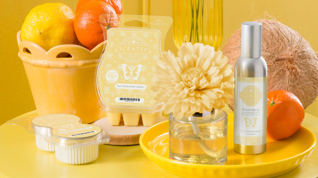 Scentsy Sun-Drenched Cotton Pods, Wax Bar, Fragrance Flower, Room Spray, a coconut, and an orange all decorate a bright yellow tabletop with matching bowls, cups, and plates.