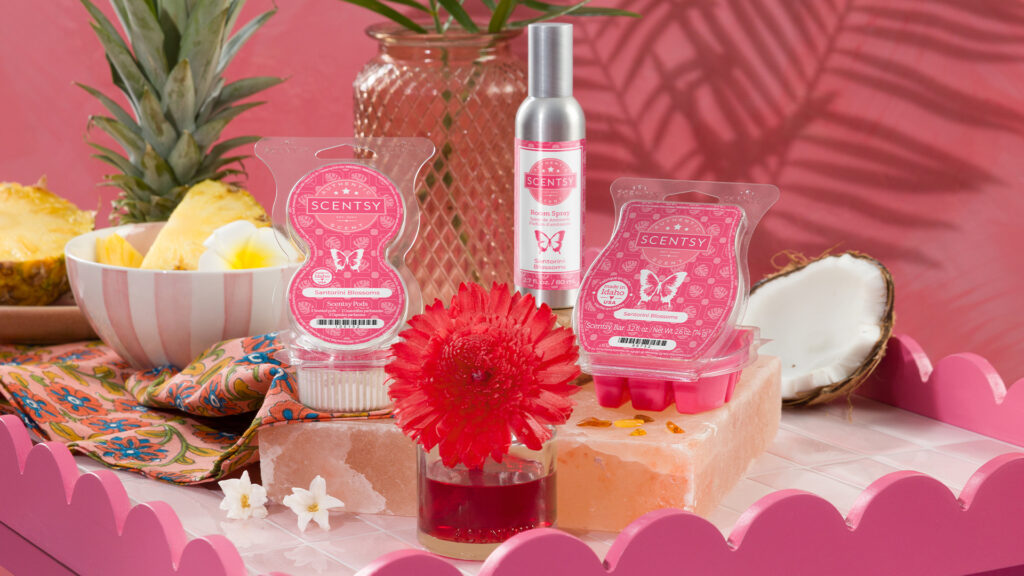Scentsy Santorini Blossoms Pods, Fragrance Flower, Room Spray, Wax Bar, a coconut, and a pineapple all decorate a pink tabletop with matching bowls, vases, and other decorations.
