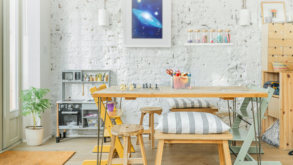 A modern art room and project space with wooden and metal chairs, table, and benches. A chess board sits on the table beside a bucket of craft tools. A white brick wall in the background is adorned with shelves filled with nicknacks and space-themed wall art. 