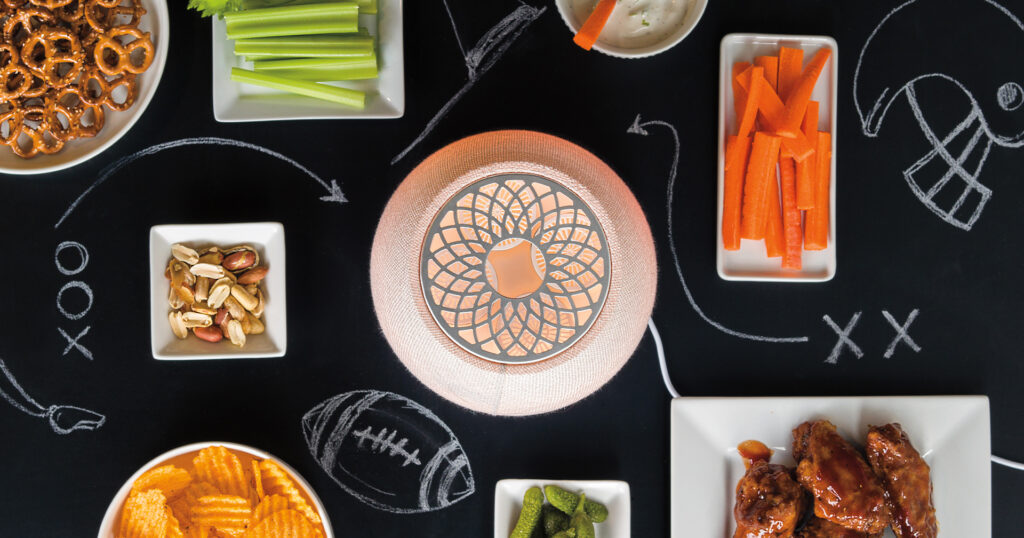 The Scentsy Air Quad Diffuser sits in the center of a blackboard surrounded by game day snacks like wings, carrots and dip, chips, and pretzles. Chalk drawings in the style of football plays surround the different dishes and Scentsy Product.