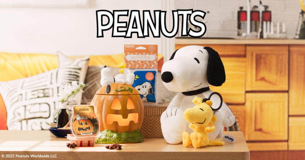 A Snoopy Scentsy Buddy stuffed toy sits beside a smaller Woodstock  Buddy Clip stuffed toy on a wooden coffee table. Beside them are the Snoopy's Pumpkin Patch Scentsy Warmer, which depicts a large pumpkin with Snoopy sleeping on top, as well as the Snoopy's Autumn Adventure Wax Bar open beside the warmer. This Scentsy Peanuts collection sits in a warmly lit room with a yellow couch and paneled cabinets in the background. 