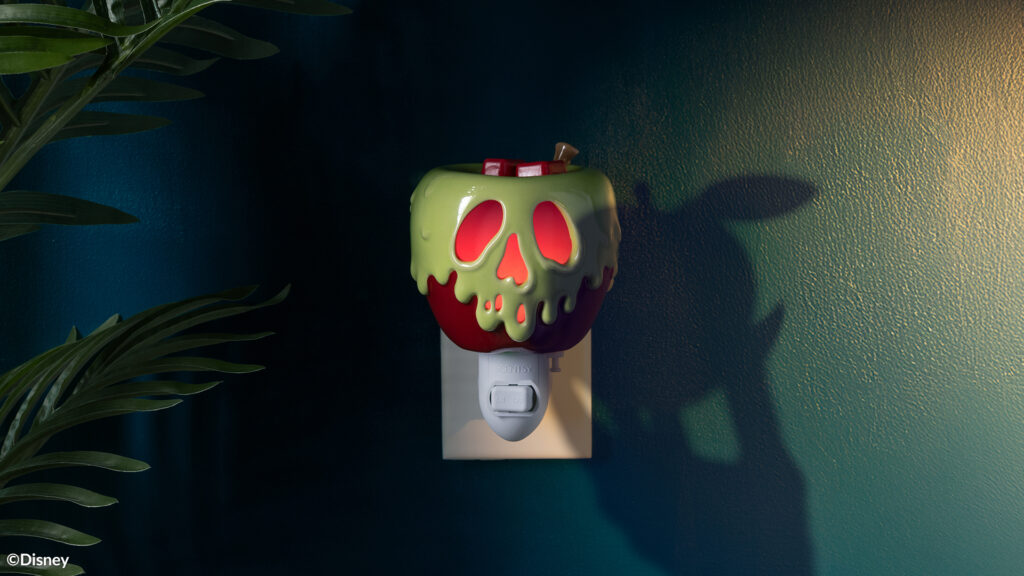The Disney Villains: Just One Bite Scentsy Mini-Warmer, shaped to look like an apple with deathly poison dripping from it like the apple from Snow White, is plugged into a wall outlet. The shadow of a hand holding an apple is cast on the wall beside the mini-warmer.