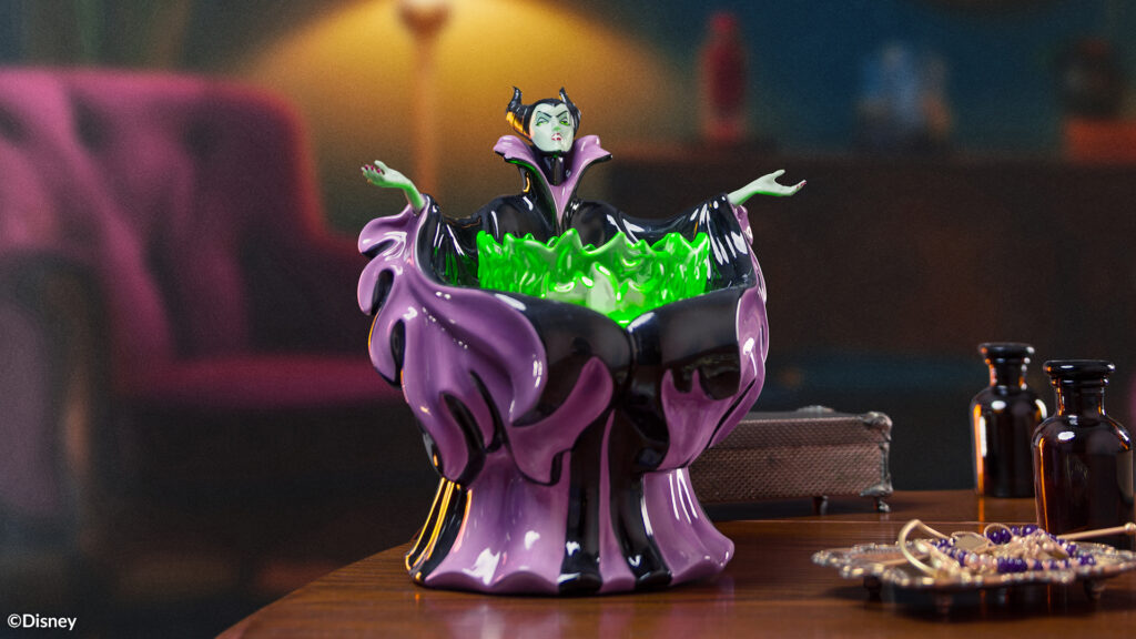 The Disney Villains: Made for Mayhem Scentsy Warmer sits on a side table beside some glass vials and jewelry. The warmer is design to look like Maleficent from Disney's Sleeping beauty, her cloak swirling around her with green fire summoned up between outstretched arms.