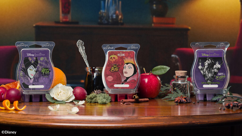 Scentsy Wax Bars Disney Maleficent: Mistress of Evil, Disney Evil Queen: just one bite, and Disney Villains: Dark and Devious are displayed on a dark wood table with apples, oranges, cherries, rose blossoms, cinnamon sticks, and vials of herbs displayed around them.