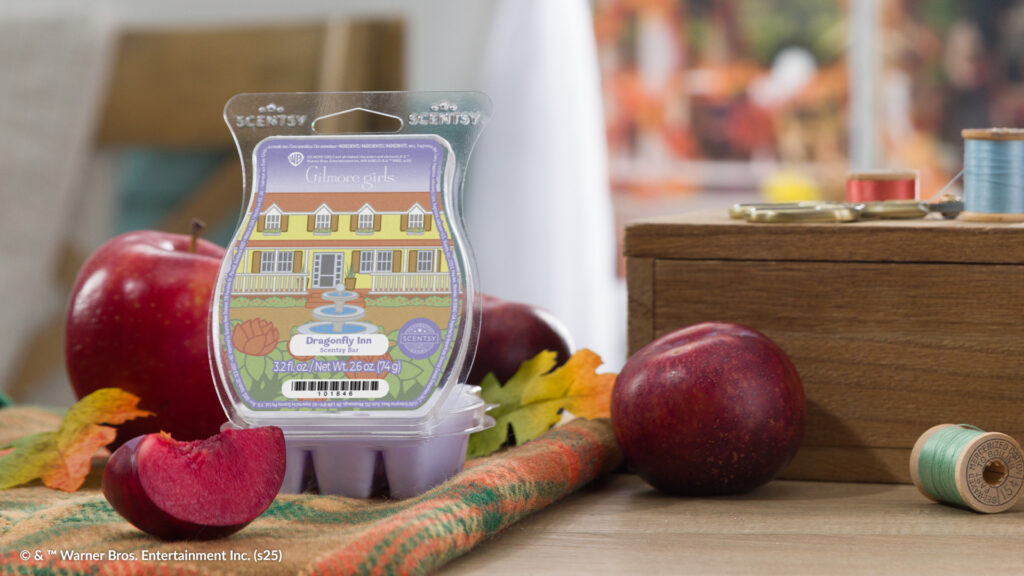 The Gilmore Girls Dragonfly Inn Scentsy Wax Bar, with lavender wax melt cubes and a label depicting a yellow hotel with white trim and a fountain surrounded by flowers, sits on a plaid blanket surrounded by plums, apples, and fallen yellow and orange leaves.