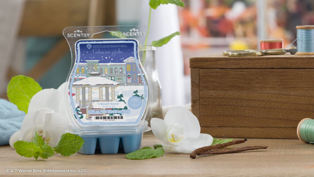 The Gilmore Girls "I Smell Snow" Wax Bar, with blue wax melt cubes and its label depicting an illustrated gazebo and quaint American street covered in snow, sits surrounded by vanilla blossoms, mint leaves, and spools of green and blue thread. 