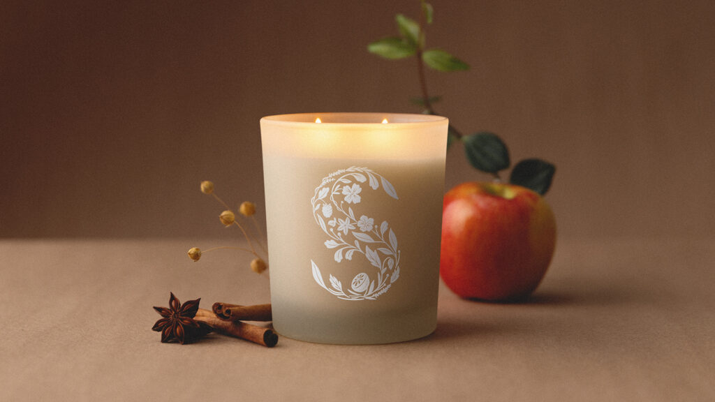 A beige colored Scentsy Candle with a white, monogrammed "S" on its frosted glass vessel sits on a light brown table beside cinnamon sticks and an apple.