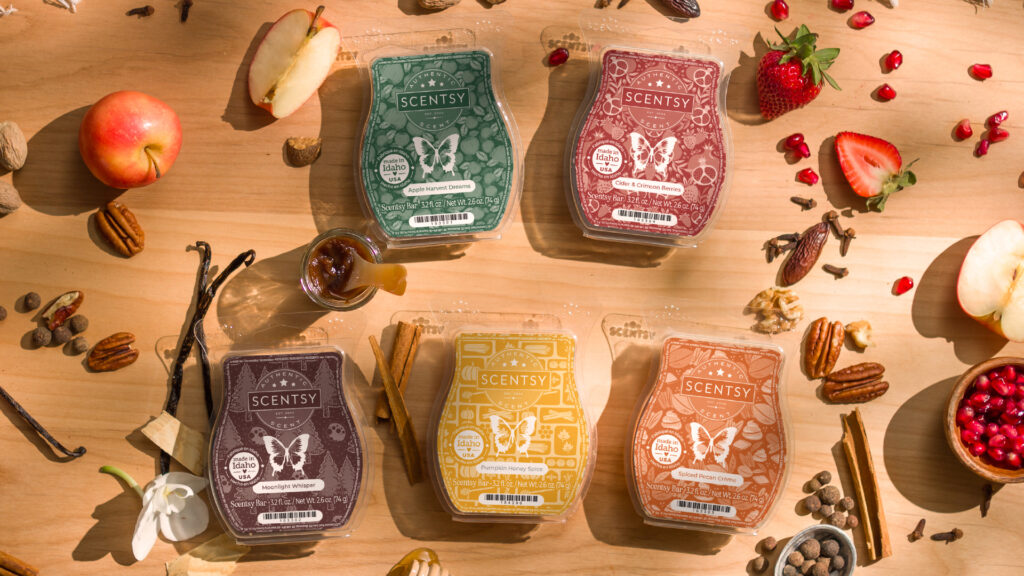 Four Scentsy Wax Bars with green, red, brown, yellow, and orange labeled wax melt clamshells sit on a wooden tabletop surrounded by walnuts, apples, cinnamon sticks, pomegranate seeds, strawberries, spices, and flower blossoms. 