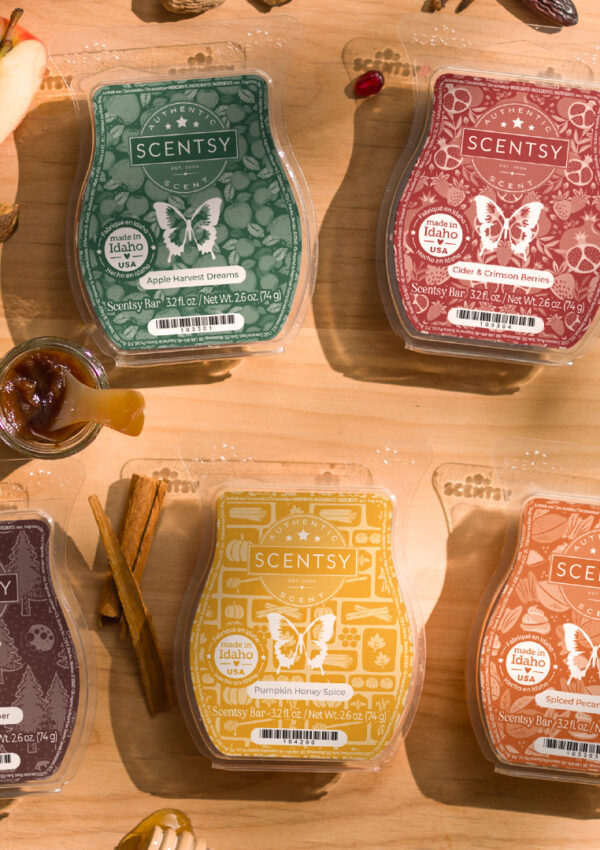 Four Scentsy Wax Bars with green, red, brown, yellow, and orange labeled wax melt clamshells sit on a wooden tabletop surrounded by walnuts, apples, cinnamon sticks, pomegranate seeds, strawberries, spices, and flower blossoms.