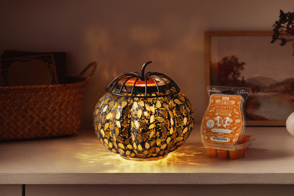 The Golden Gourde Wax Warmer, shaped like a pumpkin covered with golden leaves glowing from within, sits on a white shelf casting light across photos, a small basket, the shelving, and an orange labeled Scentsy Wax Bar clamshell full of orange wax cubes.