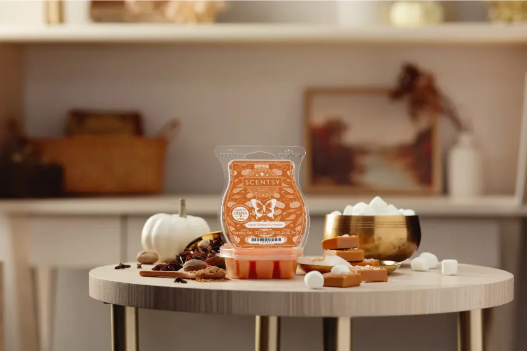 A Have S'more Pumpkin Scentsy Wax Bar wtih an orange lable sits surrounded by marshmallows, cinnamon, caramel squares, and little white pumpkins on ash-colored, circular wooden table. In the background a living room shelf with photos and flowers can be seen.
