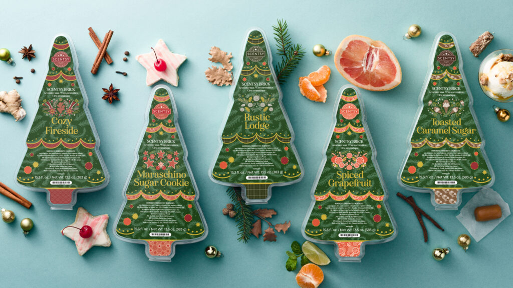 Four Christmas tree shaped Holiday Bricks from Scentsy with holiday scented wax melts sitting on a blue table surrounded cinnamon sticks, star-shaped cookies, pine needles, oranges, limes, walnuts, star anise, and ornaments - all of which represent fragrance notes and holiday cheer.