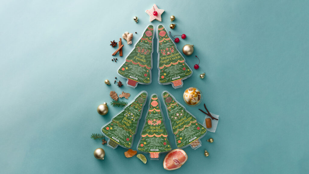 An arrangement of five Christmas tree shaped Scentsy Wax Bricks meant to depict holiday bells, all of which surrounded by cinnamon sticks, ginger root, star anise, pine needles, caramel, cherries, and other aromatics to indicate fragrance notes.
