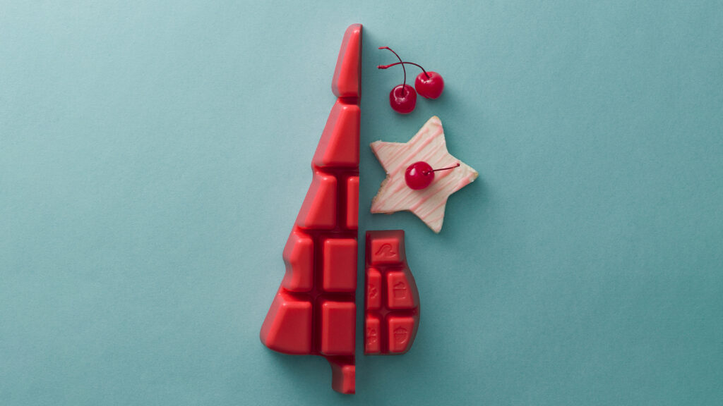 Red Scentsy wax melts cut in half and shaped into a Christmas tree, posed on a blue table beside a cookie and some cherries to indicate the fruity, sweet fragrance notes of the Wax Bar and Wax Brick. 