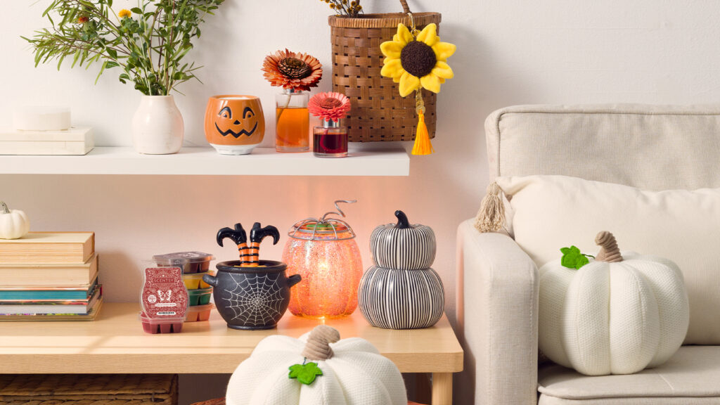 A home living room with white couch, desk, and shelving displaying Scentsy Harvest and Halloween decor, such as pumpkin shaped Warmers, cozy fragranced wax melts, Scentsy Fragrance flowers, air diffusers, buddy charm clips, and a large white pumpkin plush.