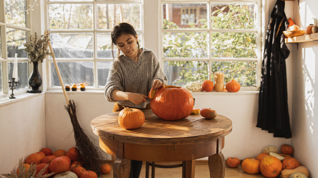 A young woman carves a pumpkin in a windowed room filled with sunshine and stacks of pumpkins.