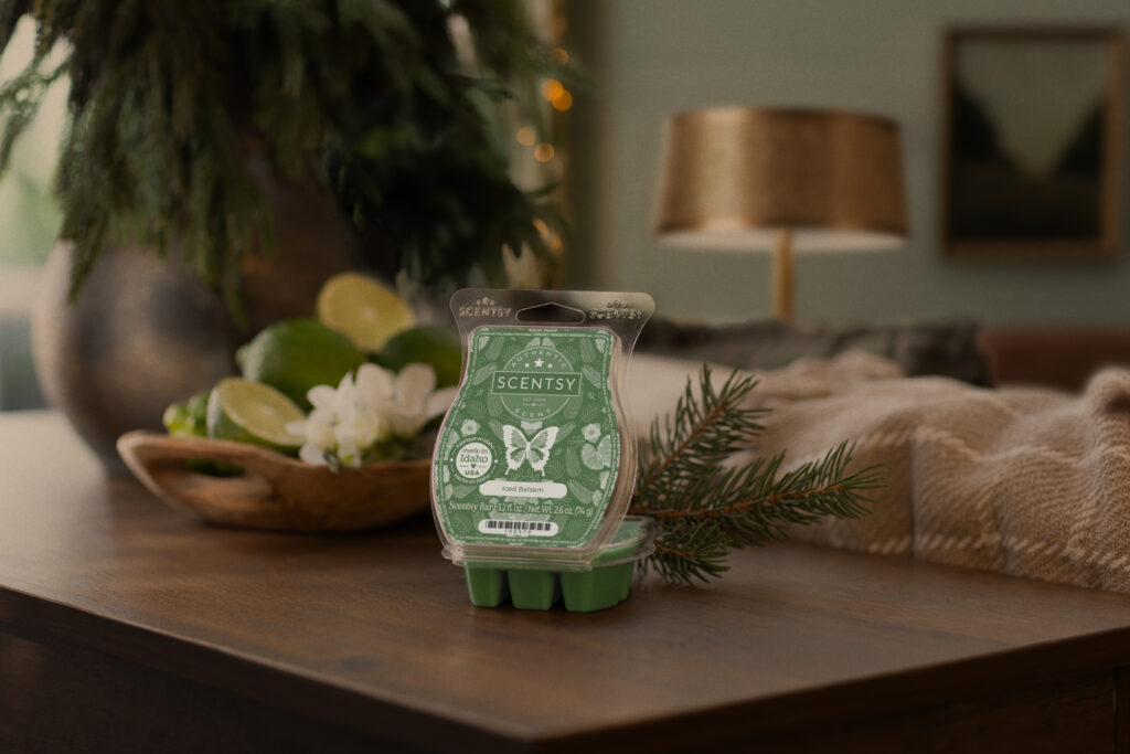 Scentsy's Scent of the Month, the Iced Balsam Wax Bar, sits on a wooden sofa table beside a fir sprig and small wooden bowl of cut limes and white freesia blossoms.