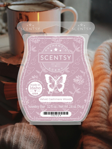 A pink labeled Scentsy wax melt in the Velvet Cashmere Woods fragrance appears overtop a scene of folded wool blankets, a cup of sweet, creamy coco, and a glowing candle.