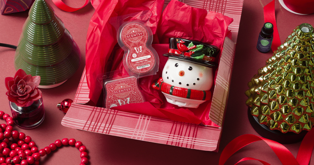 A Scentsy Gift Set Christmas gift with Scentsy warmer, wax bar, and air diffuser pods in a red box and red wrapping paper. The gift box is surrounded by red ribbons and beads, a poinsettia fragrrance flower, and Christmas tree Warmers and Diffusers.