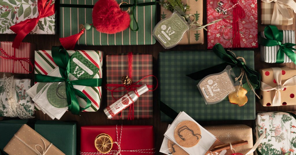 Scentsy Wax Bars, Room Sprays, Charm Clips, and Scent Circles sit on top of dozens of presents wrapped with patterned Christmas wrapping paper, ribbons, and bows. 