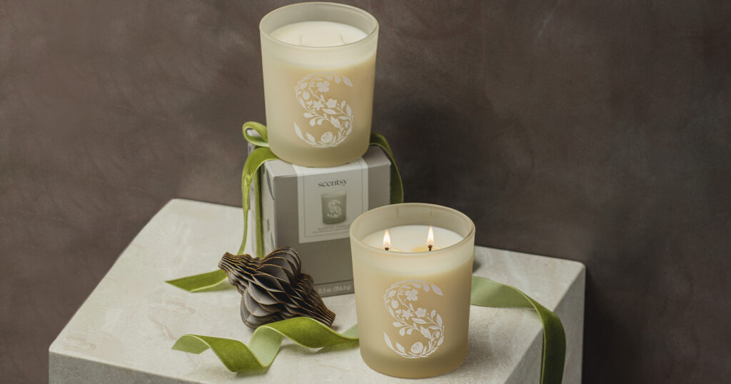 Scentsy two-wick scented candles stacked on ribbon-wrapped boxes beside Christmas decor.