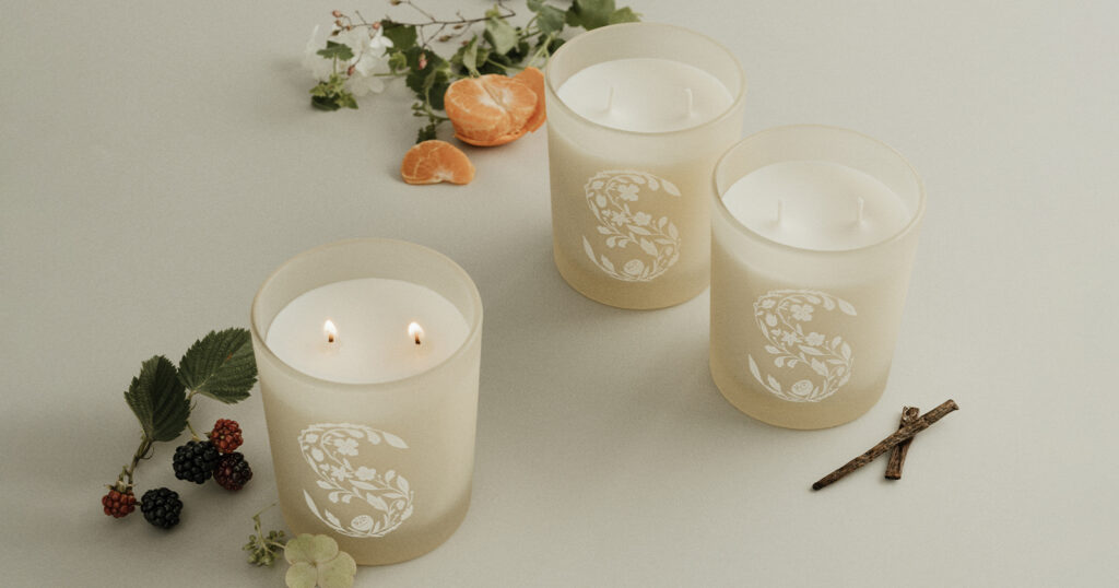 Three two-wick Scentsy scented candles sit on a white surface surrounded by orange slices, flowers, cinnamon sticks, and bergamont - each depicting the fragrance notes of the candles. 