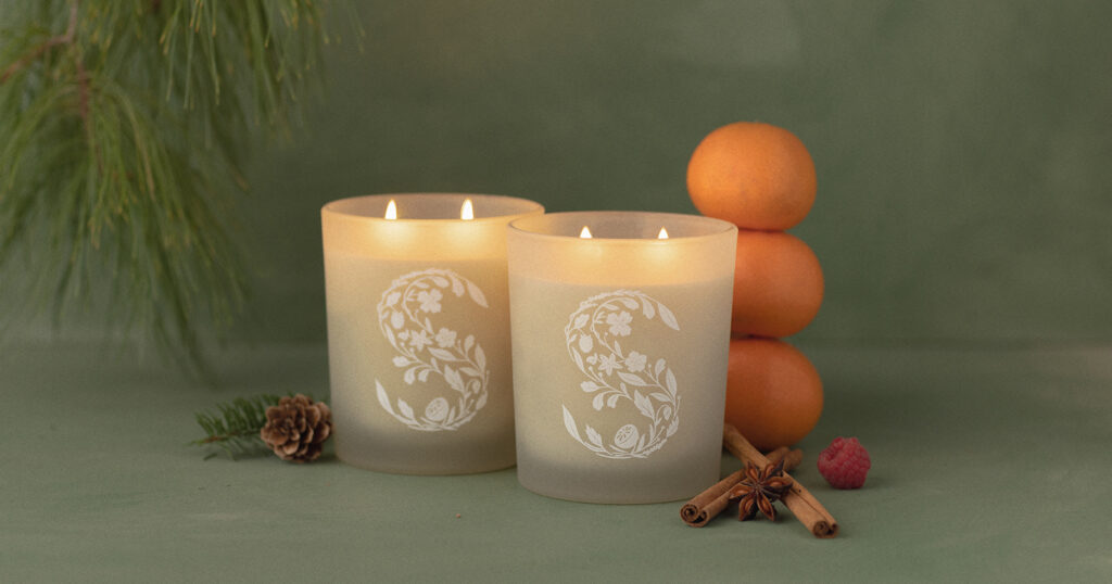 Two lit two-wick Scentsy Candles sit on a green surface beside a stack of oranges, some cinnamon sticks, pine branches and pine cones. 