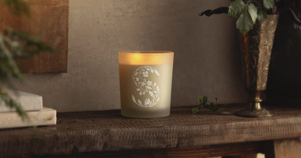 A lit Scentsy Candle sits on a dark wooden table beside house plants. 